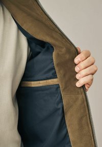 Interieur van een bruine suède jas met een blauwe voering, voorzien van een horizontale zak met een tankleurige afwerking. Ritsluiting langs de rand.