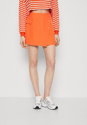 Femme portant une mini-jupe orange vif avec des boutons et un détail de poche, associée à un haut à manches longues rayé rouge et blanc et des baskets blanches.