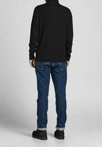 Homme portant un pull à col roulé noir, un jean bleu retroussé et des bottes noires, debout devant un fond gris clair uni.