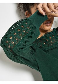 Suéter verde oscuro con intrincados detalles de encaje en las mangas, con un acabado texturizado y puños acanalados. El diseño enfatiza elementos hechos a mano.