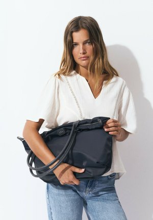 Gran bolso negro con dos correas para el hombro, fabricado en material suave. Parte superior con cremallera. El usuario lleva una blusa de color claro y jeans de mezclilla.