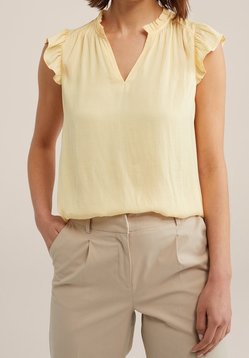 Femme portant un haut sans manches jaune clair avec des épaules froncées, rentré dans un pantalon beige, les mains dans les poches.