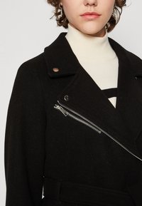 Svart ylleblandad rock med dragkedja framfickor, slagkrage och bälte i midjan. Texturen är mjuk med en skräddarsydd passform.