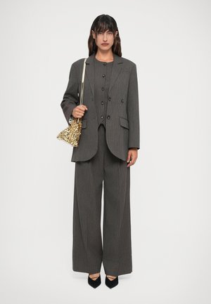 Woman wearing a grey pinstripe suit with wide-leg trousers, black pointed heels, holding a small gold sequin shoulder bag.