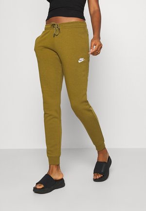 Personne portant un pantalon de survêtement Nike vert olive avec un cordon et des sandales slides noires, debout devant un fond uni gris clair.