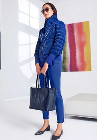 Marineblauer Pufferjacke mit hohem Kragen, kombiniert mit einem passenden Pullover und einer Hose. Eine strukturierte schwarze Tragetasche mit Monogramm. Graue Ballerinas runden das Outfit ab.