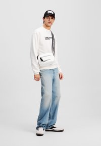 Sweatshirt bianco con testo grafico nero, jeans in denim blu chiaro e una borsa a tracolla bianca. Un cappellino nero e delle sneakers completano il look.