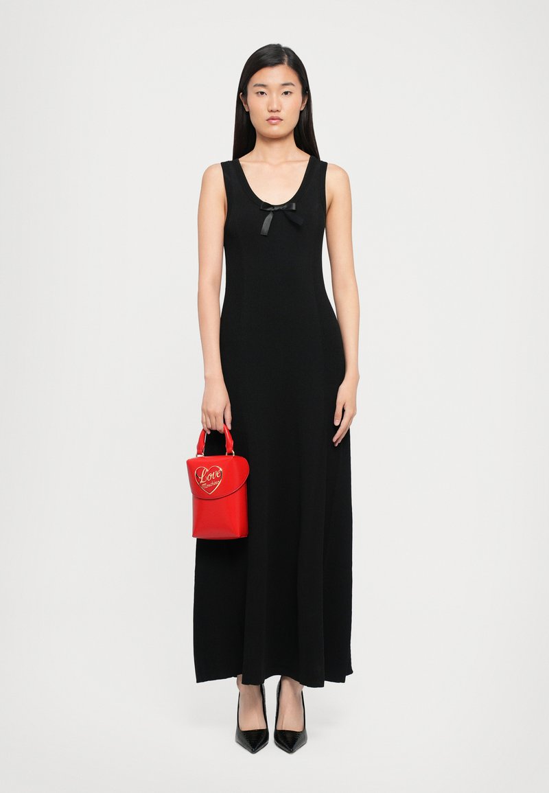 Woman in a sleeveless black maxi dress with a small black bow, holding a red handbag with gold heart-shaped "Love" detail, wearing black heels.
