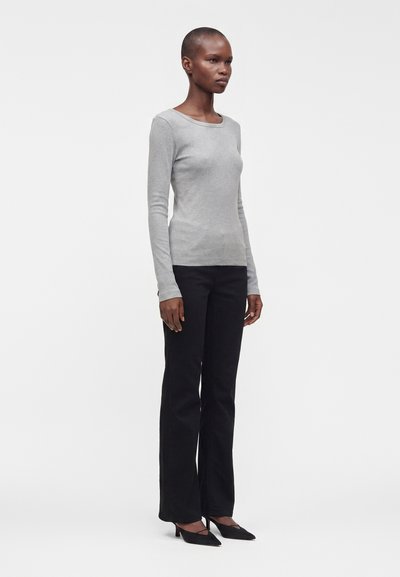 Femme aux cheveux courts portant un haut gris clair moulant à manches longues, un pantalon noir évasé et des talons noirs pointus, debout de profil.