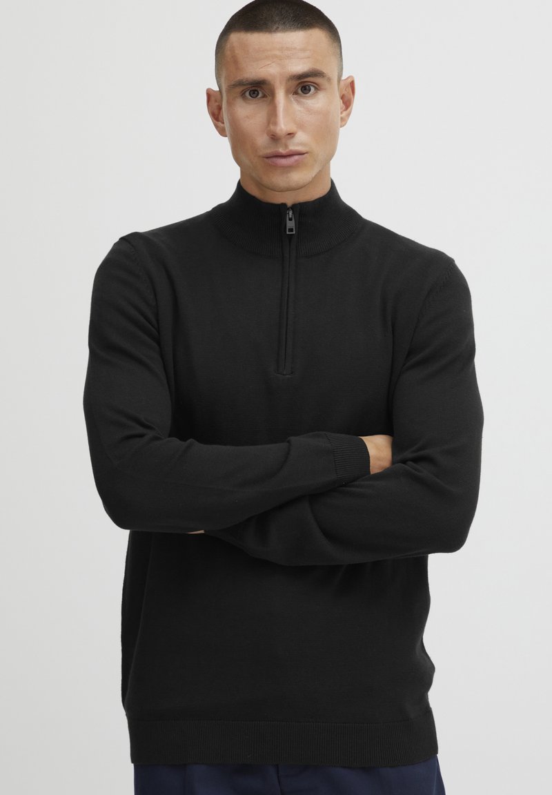 Black zip-up sweater with a high collar, long sleeves, ribbed cuffs, and textured fabric. Minimalist design with no visible patterns or logos.