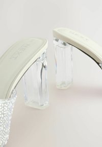 Sandales à talons blocs transparentes avec accents en perles, présentant une tige blanche lisse et un talon transparent. Le design est élégant et moderne.