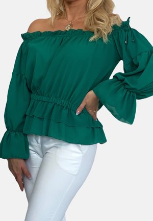 Kvinde iført grøn off-shoulder bluse med flæser på ærmerne og taljen, kombineret med hvide bukser, hånd hvilende på hoften.