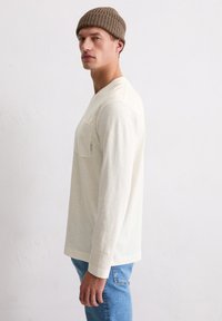 Mann, der eine beige Strickmütze, ein cremefarbenes Langarmshirt mit Fronttasche und blaue Jeans trägt, steht seitlich vor einem schlichten Hintergrund.