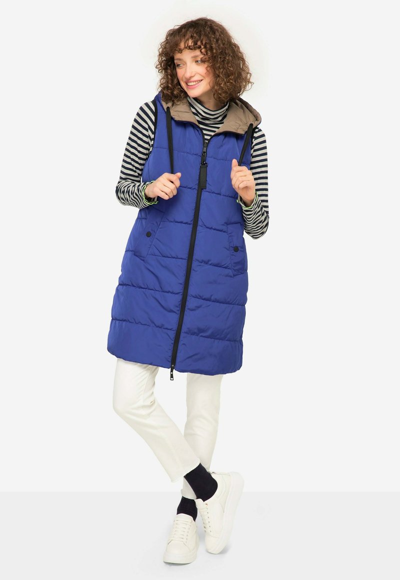 Gilet en duvet bleu avec une capuche beige, comportant une fermeture éclair à l'avant, deux poches latérales et un design matelassé. Porté par-dessus une chemise rayée et un pantalon blanc.