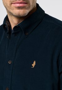 Barzdotas vyras, dėvintis tamsiai mėlyną švarką iš kantavimo audinio su mažu šviesiai ruda siuvinėta kengūros logotipu krūtinės srityje.