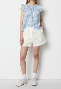 Blusenes blaues Blumenoberteil mit rüschigen Schultern, kombiniert mit weißen, hoch taillierten Shorts und weißen Sneakers mit lila Akzenten.