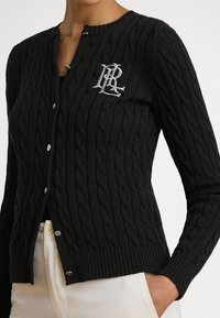 Μ�αύρη ζακέτα με πλέξη καλωδίου και ασημένιο λογότυπο, με στρογγυλή λαιμόκοψη, κούμπωμα με κουμπιά και ριμπ τελείωμα. Το υφασμάτινο υφή προσθέτει διάσταση.