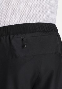Pantalones cortos atléticos negros con cintura elástica, con un bolsillo trasero y detalle de cordón. El material parece suave y ligero.