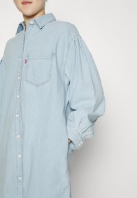Camicia di denim azzurro chiaro con design a bottoni, tasca sul petto e polsini arrotondati. Presenta una vestibilità ampia e una consistenza morbida.