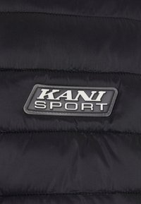 Melnas pūšu jakas ar KANI SPORT loga uzšuvumu. Materiāls ir gluds, ar izšūtām quilted rakstainēm visā virsmā.