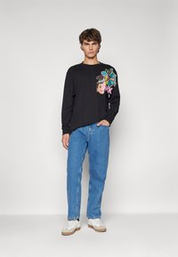 Camicetta nera a maniche lunghe con stampe grafiche colorate sul davanti. Abbinata a jeans di denim blu chiaro e sneakers bianche con dettagli beige.