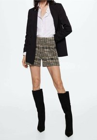 Blazer doppiopetto nero, camicia bianca con bottoni, pantaloni corti a quadri in beige e marrone, stivali neri in suede al ginocchio con punta quadrata.