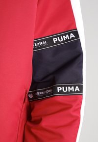 Manga de chaqueta roja con rayas en blanco y negro, con la marca "PUMA" y el texto "INTERNATIONAL FORTY EIGHT". Textura de tela suave.
