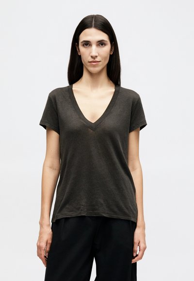 Junge Frau mit glatten, dunklen Haaren, die ein dunkelgraues, kurzärmeliges Shirt mit V-Ausschnitt und schwarze Hosen trägt, steht vor einem schlichten weißen Hintergrund.