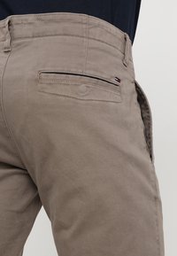 Beżowe bawełniane spodnie z tylną kieszenią z czarnym zamkiem błyskawicznym oraz małym logo w kolorach czerwonym, białym i niebieskim nad kieszenią.