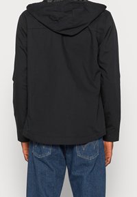 Giacca nera con cappuccio dal design semplice, caratterizzata da una texture liscia e da un taglio dritto all'orlo. Indossata con pantaloni in denim blu.