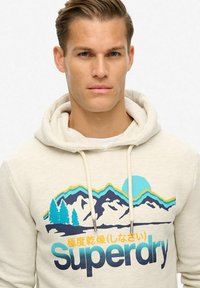 Beżowy hoodie z graficznym wzorem przedstawiającym góry, drzewa i słońce w niebieskim, zielonym i żółtym kolorze, z regulowanymi sznurkami i przednią kieszenią.