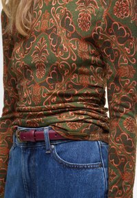 Lange mouwen shirt in donkergroen met intrikate oranje bloemen- en ananaspatronen; gecombineerd met blauwe denim jeans en een bordeauxrode riem.