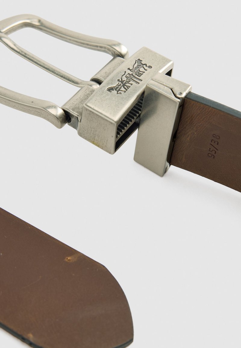 Brun læderbælte med en metallisk sølvspænde, der har et logo og en tekstureret fastgørelsespunkt, med detaljerede syninger langs kanterne.