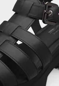 Κοντινό πλάνο μαύρου δερμάτινου σανδαλιού με τρεις οριζόντιες λουράκια και λαμπερό μεταλλικό πόρπη σε χοντρή σόλα.