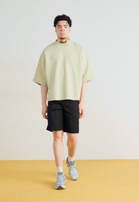 Camisa de manga corta oversized verde claro, pantalones cortos negros, zapatillas grises, de pie sobre un suelo amarillo, contra un fondo liso.