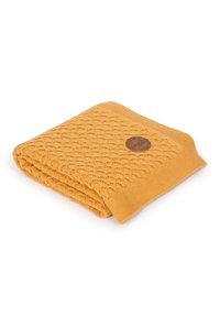 Żółty dziany koc z wzorem fal, wykończony prążkowanym brzegiem, z okrąłą brązową metką w jednym rogu.