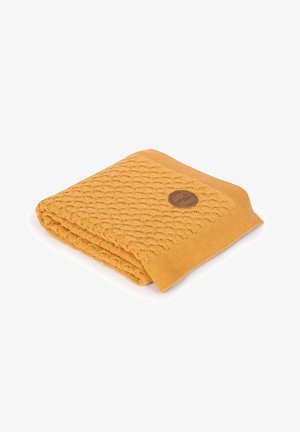 Żółty dziany koc z wzorem fal, wykończony prążkowanym brzegiem, z okrąłą brązową metką w jednym rogu.