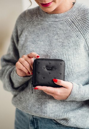 Black leather wallet with embossed dot pattern, zip closure, and logo. Held by a person wearing a gray sweater. Red nails are visible.