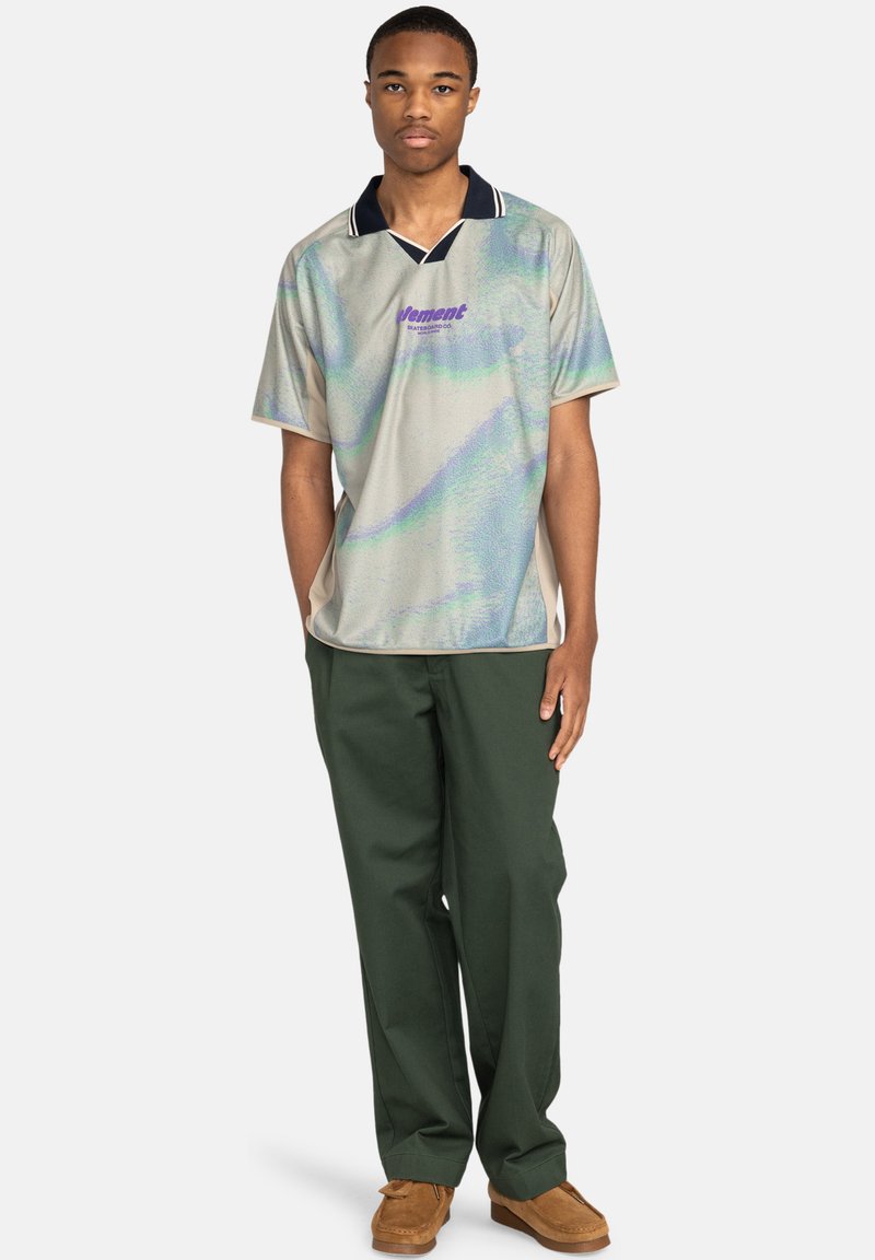 Poloshirt mit kurzen Ärmeln aus schimmerndem Stoff in Silber- und Türkistönen, ausgestattet mit einem marineblauen Kragen und einem lila Logo auf der Brust, kombiniert mit grünen Hosen.