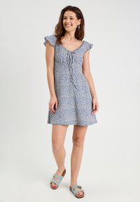 Light blue dress with white floral pattern, flared shape, short ruffled sleeves, and a tie front detail. Paired with blue sandals.
