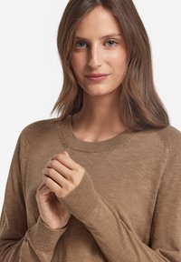 Brun stickad tröja med rund halsringning, ribbade ärmar och en slät textur; modellens händer är försiktigt hopknäppta fram.