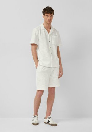 Mann steht, trägt ein strukturiertes weißes kurzärmeliges Hemd mit Knöpfen und passende kniehohe Shorts mit weißen Sneakern.