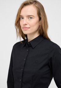 Black button-up shirt features a classic collar, front placket, and subtle logo embroidery on the chest; smooth fabric with a structured fit.