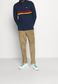 Marinblå långärmad polo med röda och gula horisontella ränder, ihop med beige kordflorsbyxor och vita sneakers med gröna snören.