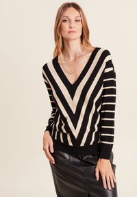 V-Ausschnitt Pullover mit vertikalen und diagonalen schwarzen und beigen Streifen. Weicher Stoff mit einer taillierten Silhouette. Kombiniert mit einem schwarzen Lederrock.
