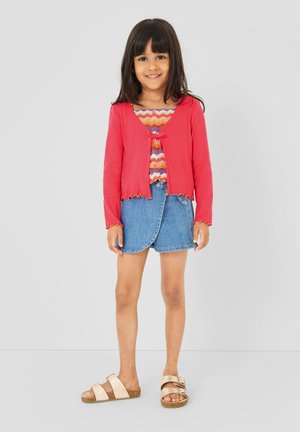 Młoda dziewczyna uśmiechająca się, ubrana w czerwony kardigan na guziki, wielokolorowy wzorzysty top, niebieską spódnicę dżinsową oraz beżowe sandały.