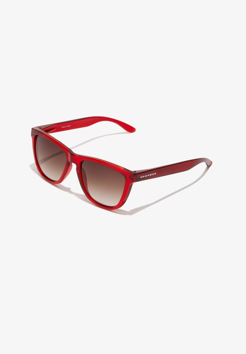 Gafas de sol de plástico transparente rojo con un marco ancho, lentes con tinte marrón gradual y la marca "HAWKERS" impresa en la patilla.