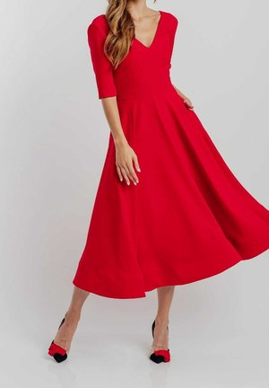 Donna che indossa un vestito rosso di lunghezza media con maniche tre quarti e tacchi neri con fiocchi rossi, in piedi davanti a uno sfondo chiaro e semplice.