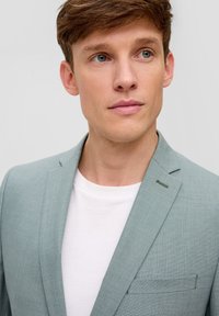 Helles grünes Blazer mit strukturiertem Stoff, einzelner Reverskragen und Akzent auf der Brusttasche. Über einem weißen T-Shirt getragen.