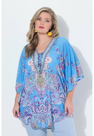 Kvinde med langt blondt hår iført gule blomsterøreringe og en løs blå top med indviklede blomstermønstre og paisley-designs.